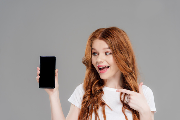 schöne aufgeregte rothaarige Mädchen zeigt mit dem Finger auf Smartphone mit leerem Bildschirm isoliert auf grau - Foto, Bild