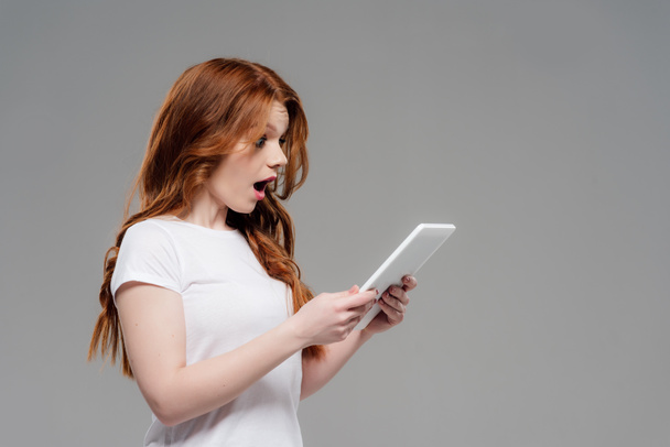 mooie verrast roodharige meisje met behulp van digitale Tablet geïsoleerd op grijs - Foto, afbeelding