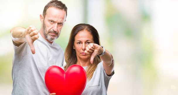 Středním věku hispánský příležitostné pár v lásce držící červené srdce nad izolované pozadí s naštvaný obličej, záporné znaménko ukazující nechuť s palci dolů, koncept odmítnutí - Fotografie, Obrázek