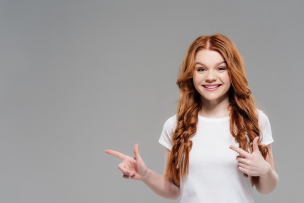 beautiful redhead girl looking at camera, smiling and pointing with fingers isolated on grey with copy space - Φωτογραφία, εικόνα