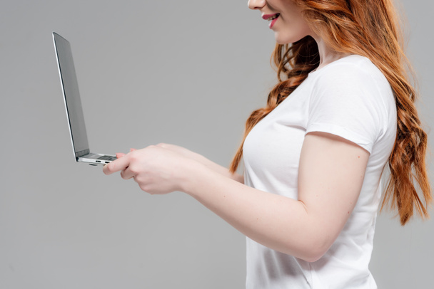 oříznutý pohled na zrzanou dívku v bílém tričku s notebookem izolovaným na šedé - Fotografie, Obrázek