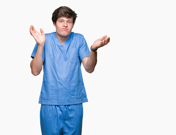 Giovane medico indossa uniforme medica su sfondo isolato espressione indiscreta e confusa con le braccia e le mani sollevate. Concetto di dubbio
. - Foto, immagini