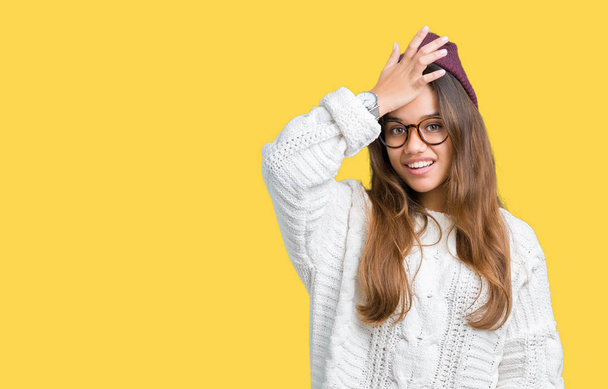 Jonge mooie brunette hipster vrouw dragen van bril en winter muts over geïsoleerde achtergrond verrast met de hand op het hoofd voor fout, niet vergeten fout. Vergat, slecht geheugen concept. - Foto, afbeelding