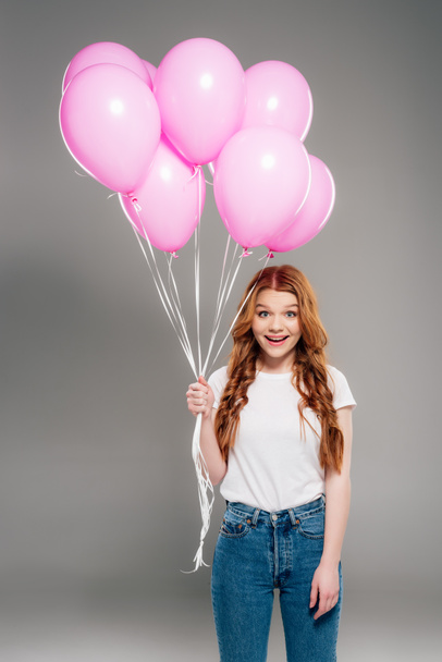beautiful surprised girl with red hair holding pink air balloons and looking at camera on grey - 写真・画像