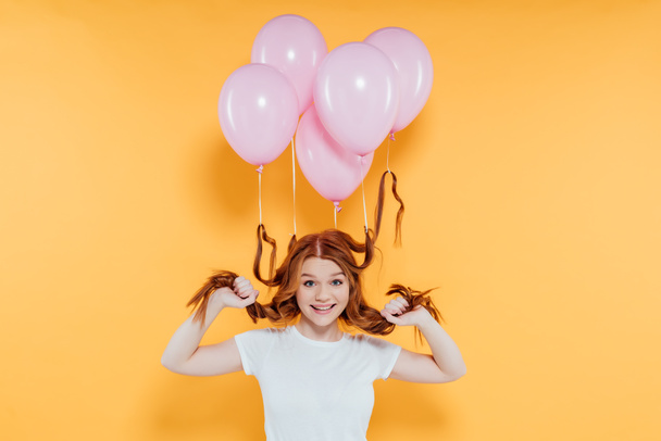 happy redhead girl with balloons tied to hair posing isolated on yellow - Φωτογραφία, εικόνα