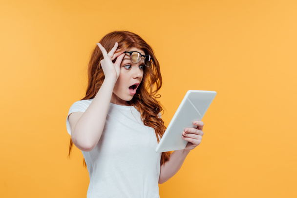surprised redhead girl with glasses using digital tablet isolated on yellow - Photo, Image