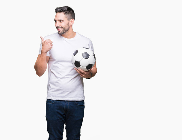 Νέος άνθρωπος που κρατά την μπάλα ποδοσφαίρου ποδοσφαίρου πέρα από το απομονωμένο υπόβαθρο κατάδειξης και δείχνοντας με αντίχειρα μέχρι την πλευρά με το χαρούμενο πρόσωπο χαμογελά - Φωτογραφία, εικόνα