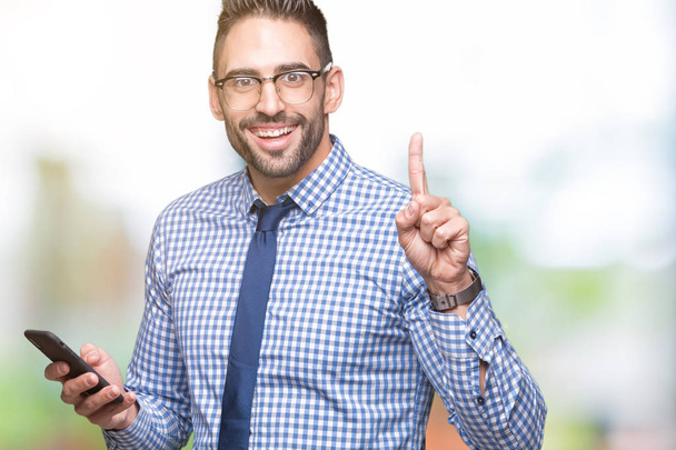 Jonge zakenman met smartphone via geïsoleerd achtergrond verrast met een idee of vraag wijzende vinger met blij gezicht, nummer één - Foto, afbeelding