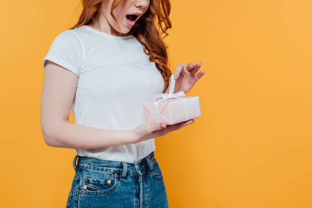 cropped view of surprised redhead girl holding gift box isolated on yellow with copy space - 写真・画像