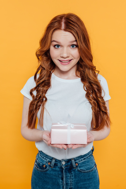 beautiful redhead girl looking at camera and holding gift box isolated on yellow - Foto, Imagem