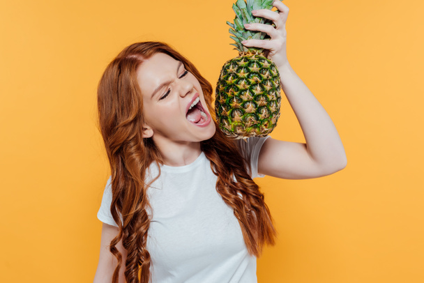 beautiful redhead girl making face expression while biting pineapple isolated on yellow - 写真・画像