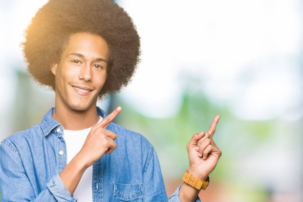 Jovem afro-americano com cabelo afro sorrindo e olhando para a câmera apontando com duas mãos e dedos para o lado
. - Foto, Imagem