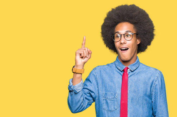 Jonge Afro-Amerikaanse zakenman met afro haar dragen van bril en rood vastbinden wijzende vinger met succesvolle idee. Verlaten en gelukkig. Nummer één. - Foto, afbeelding