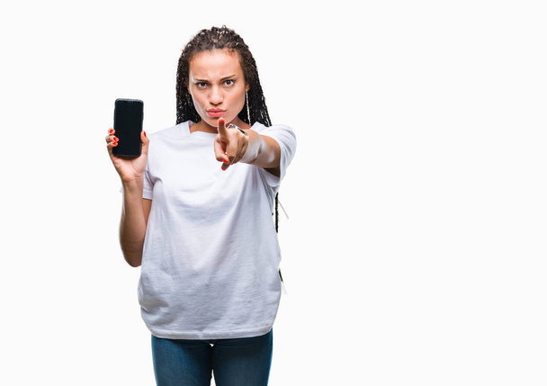 Young braided hair african american girl showing screen of smartphone over isolated background pointing with finger to the camera and to you, hand sign, positive and confident gesture from the front - 写真・画像