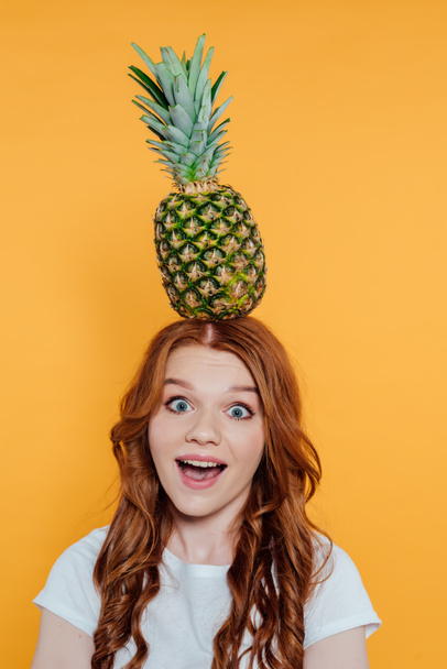 izgatott vöröshajú lány nézi a kamerát, és pózol az ananász a fejét izolált sárga - Fotó, kép