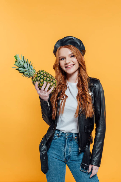 beautiful smiling redhead girl in leather jacket holding pineapple and looking at camera isolated on yellow - Foto, Imagem