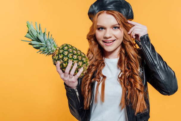 beautiful smiling redhead girl in leather jacket holding pineapple and looking at camera isolated on yellow - Фото, зображення