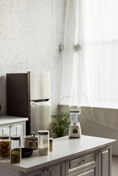Szklane pojemniki ze zbożem w pobliżu blendera na białej powierzchni w kuchni  - Zdjęcie, obraz