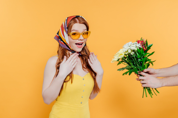 έκπληκτος κορίτσι με μαντίλα και τα γυαλιά ηλίου χειρονομία, ενώ ο άνθρωπος κρατώντας λουλούδια στο χέρι απομονώθηκαν σε κίτρινο - Φωτογραφία, εικόνα
