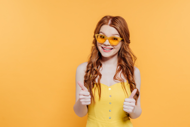 beautiful redhead girl in sunglasses looking at camera and showing thumbs up isolated on yellow - Foto, afbeelding