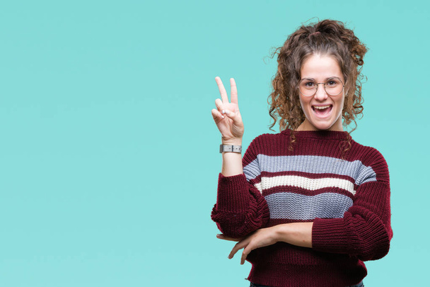 Beautiful brunette curly hair young girl wearing glasses over isolated background smiling with happy face winking at the camera doing victory sign. Number two. - Photo, Image