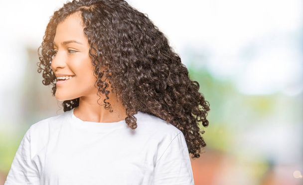 Young beautiful woman with curly hair wearing white t-shirt looking away to side with smile on face, natural expression. Laughing confident. - Foto, Bild