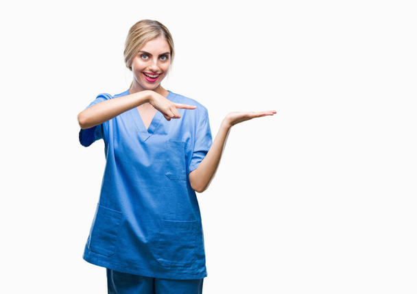 Nuori kaunis blondi lääkäri kirurgi sairaanhoitaja nainen eristetty tausta hämmästynyt ja hymyillen kameralle samalla esittää käsi ja osoittaa sormella
. - Valokuva, kuva