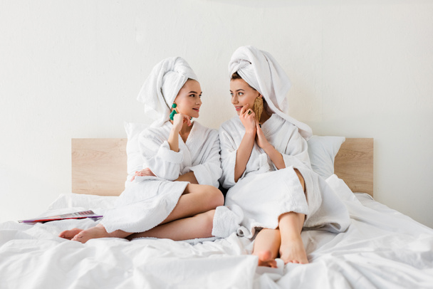 stylish girls in earrings, bathrobes and with towels on heads sitting on bed and looking at each other - Фото, изображение