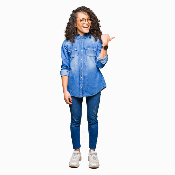 Young beautiful woman with curly hair wearing glasses smiling with happy face looking and pointing to the side with thumb up. - 写真・画像