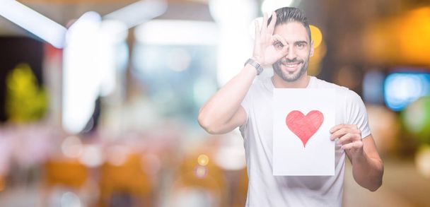 Красивый молодой человек держит карточку с красным сердцем на изолированном фоне со счастливым лицом улыбается делает хорошо знак с рукой на глазу глядя сквозь пальцы
 - Фото, изображение
