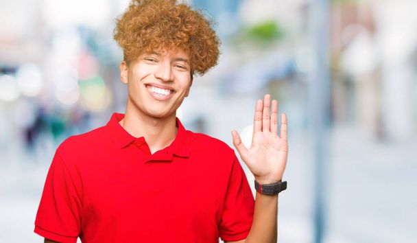 Красивий зображений юнаком iз афро волосся, що носіння червоного Waiving t-Shirt привітатися щасливі і доброзичливий, усміхнений Ласкаво просимо жест - Фото, зображення
