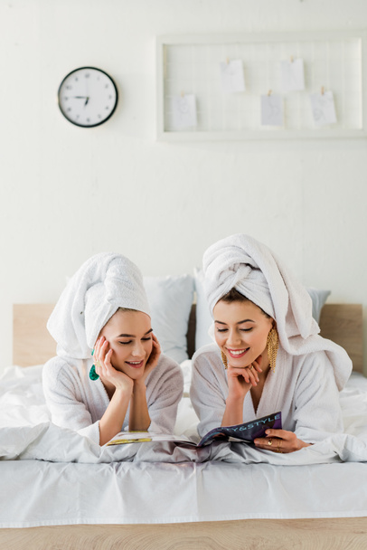 heureux élégantes femmes en peignoirs, boucles d'oreilles et avec des serviettes sur la tête lecture magazine tout en étant couché dans le lit
 - Photo, image