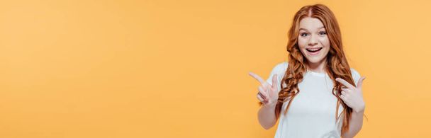 panoramic shot of beautiful smiling redhead girl looking at camera and pointing with fingers isolated on yellow - Zdjęcie, obraz