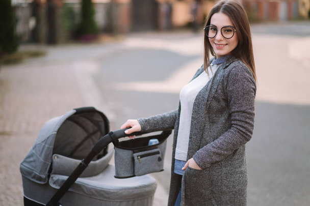Family, child and parenthood concept - Happy mother with stroller in the park. Mom with sunglasses and stylish grey jacket - Valokuva, kuva