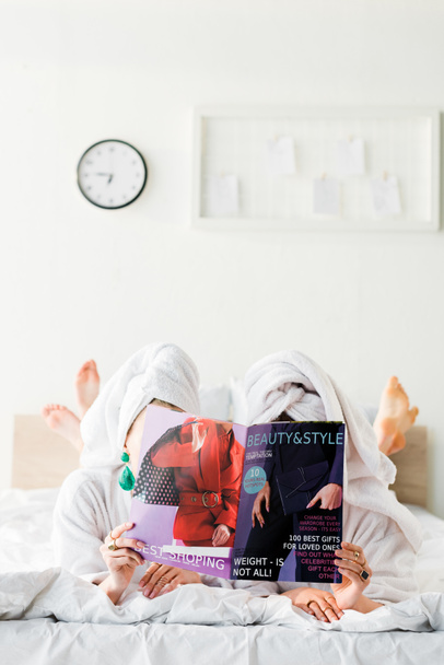 mujeres descalzas en albornoces y joyas, con toallas en las cabezas escondidas detrás de la revista mientras yacen en la cama
 - Foto, imagen
