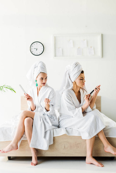 barefoot women in bathrobes and jewelry, with towels on heads using nail files while sitting on bed - Фото, зображення