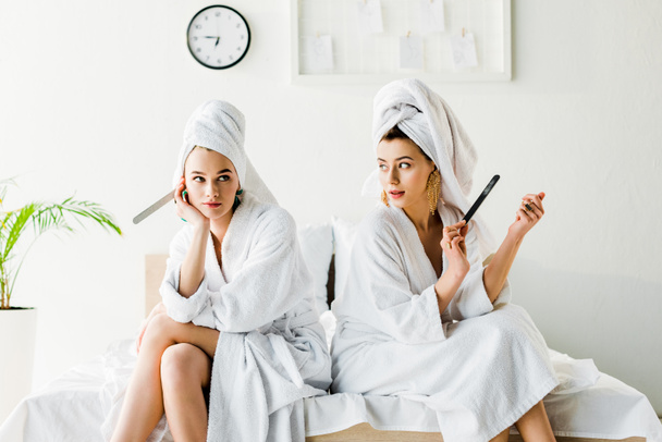 stylish women in bathrobes and jewelry, with towels on heads sitting on bed with nail files - 写真・画像