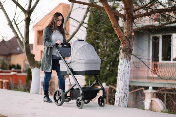 Jovem mãe andando com carrinho no parque. Mãe elegante com bebê
 - Foto, Imagem