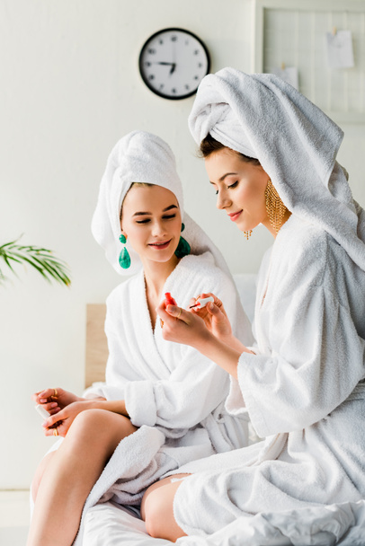 smiling stylish woman in bathrobe and jewelry, with towel on head sitting on bed and looking at friend with nail polish - Photo, Image
