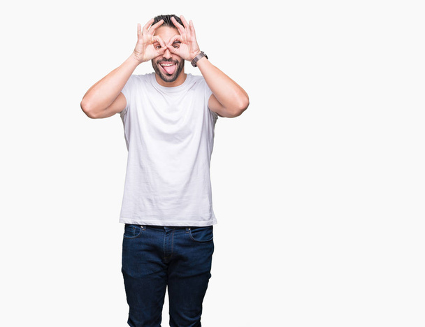 Νεαρός άνδρας φορώντας περιστασιακή λευκό t-shirt πάνω από το απομονωμένο φόντο κάνει εντάξει χειρονομία όπως κιάλια να κολλήσει τη γλώσσα έξω, μάτια αναζητούν μέσα από τα δάχτυλα. Τρελό έκφραση. - Φωτογραφία, εικόνα