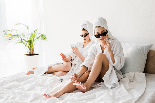 stijlvolle vrouwen in badjassen en zonnebrillen, handdoeken en sieraden liggend in bed, pedicure, roken en het gebruik van smartphone - Foto, afbeelding
