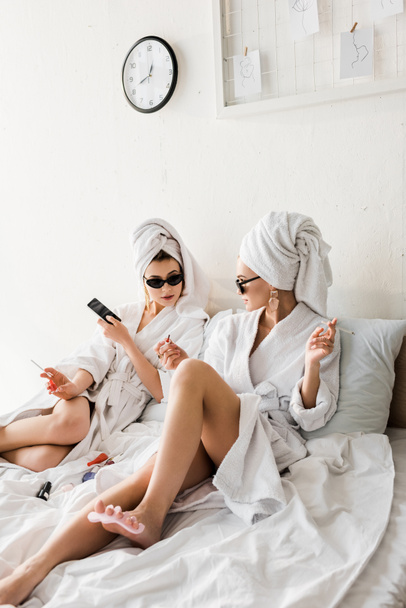 femmes élégantes en peignoirs et lunettes de soleil, serviettes et bijoux couchés dans le lit, faire pédicure et fumer
 - Photo, image