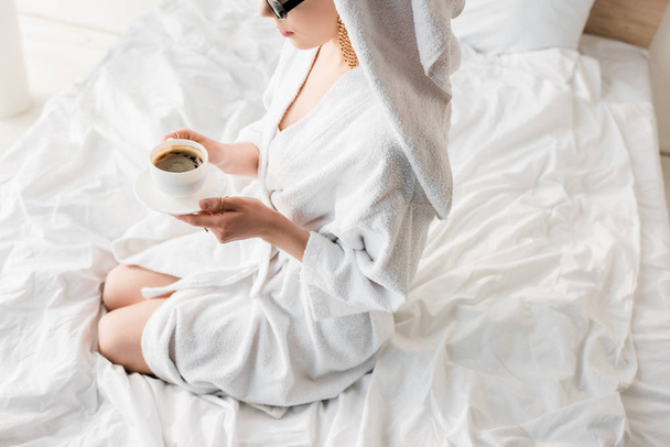 vista ritagliata di donna elegante in accappatoio e occhiali da sole, asciugamano e gioielli bere caffè a letto
 - Foto, immagini