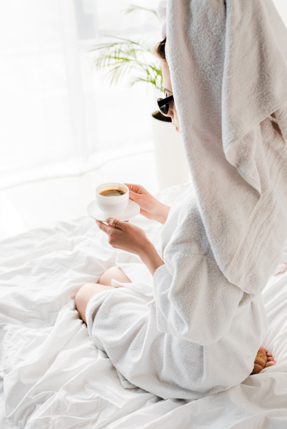 stílusos nő fürdőköpeny és napszemüveg, törölköző és ékszer gazdaság csésze kávé az ágyban - Fotó, kép