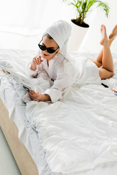 κομψό χαμόγελο ξυπόλυτη γυναίκα σε πουκάμισο, γυαλιά ηλίου, κοσμήματα και με πετσέτα στο κεφάλι διαβάζοντας εφημερίδα στο κρεβάτι - Φωτογραφία, εικόνα