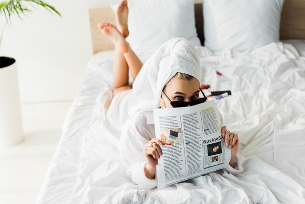 stylish barefoot woman in shirt, sunglasses, jewelry and with towel on head reading business newspaper in bed - Photo, Image