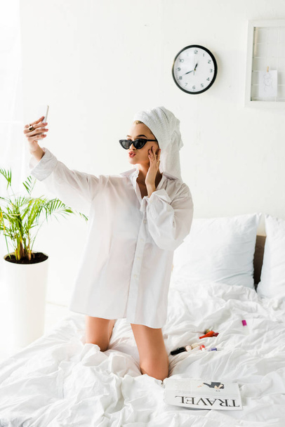 stylish woman in shirt, sunglasses, jewelry and with towel on head taking selfie in bed - Fotó, kép