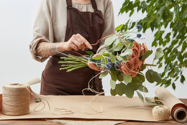 Floral design studio, woman's hands with tatto are making decorative bouquet from fresh natural fragrant flowers roses living coral color on a gray background . Concept of flower shop. - Foto, imagen