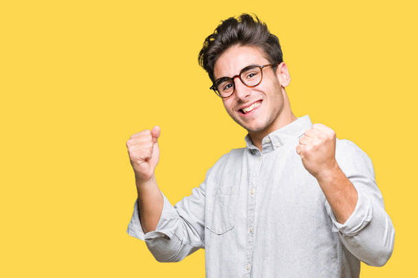 Mladý pohledný muž nosí brýle izolované pozadí velmi šťastný a nadšený, dělá vítěze gesto rukama zvedl, usmíval se a křičí na úspěch. Koncept oslav. - Fotografie, Obrázek