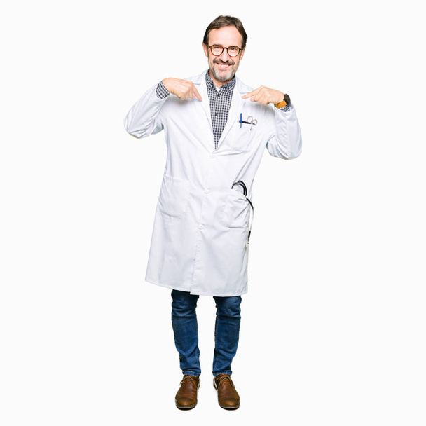 Μέση ηλικία άνδρες γιατρός φοράει ιατρική παλτό αναζητούν αυτοπεποίθηση με χαμόγελο στο πρόσωπο, δείχνοντας τον εαυτό του με τα δάχτυλα, περήφανος και χαρούμενος. - Φωτογραφία, εικόνα